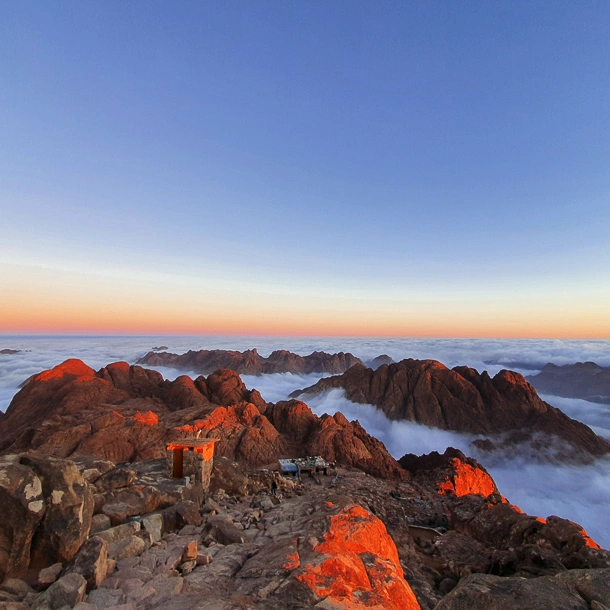 Mount Sinai summit at sunrise with clouds below, showing a golden sky and high-altitude beauty in Egypt’s mountains.