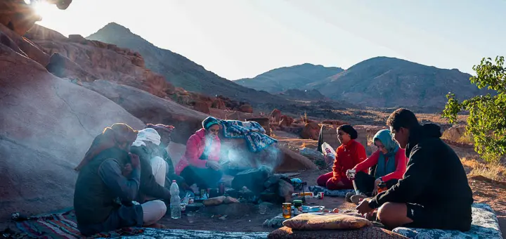 A group of hikers sits around a small fire in the early morning light, sipping tea and enjoying a simple breakfast amid the rugged mountain landscape of Saint Catherine.