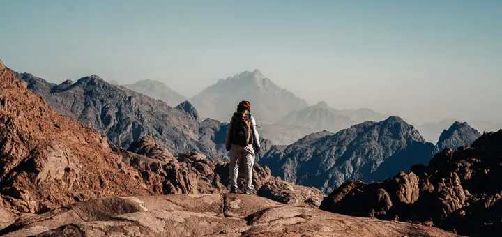 A Bedouin guide stands on a high ridge along the Bab El-Donia trail, gazing out over the distant, towering silhouette of Mount Umm Shomat under a wide Sinai sky.
