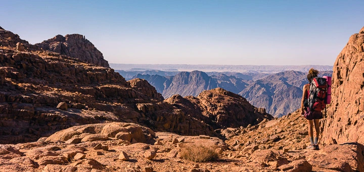 A lone hiker pauses on a rocky peak in the Saint Catherine mountains, surrounded by dramatic granite cliffs and sweeping views of the Sinai highlands under a vast open sky.
