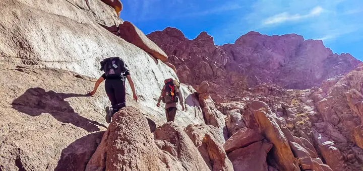 A hiker accompanied by a Bedouin guide climbs a rocky trail through Wadi Ahmar toward the summit of Mount Catherine, with golden light illuminating the granite landscape and steep mountain slopes.
