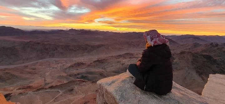 Top 10 things to do in Sinai in 2025 A Bedouin guide, dressed in traditional clothing, sits peacefully on a rocky ledge near the summit of Mount Sinai at dawn, overlooking a vast mountain range as the first light of sunrise begins to glow on the horizon.