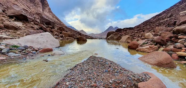 Stony trail on Mount Catherine with muddy, pooled water from a recent flash flood, under a cloudy sky.