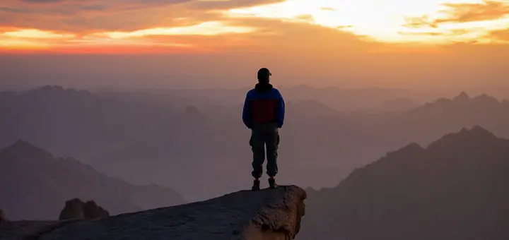 A hiker standing on Mount Sinai at sunrise, overlooking South Sinai’s mountain range bathed in golden morning light. From Egypt Mountains.